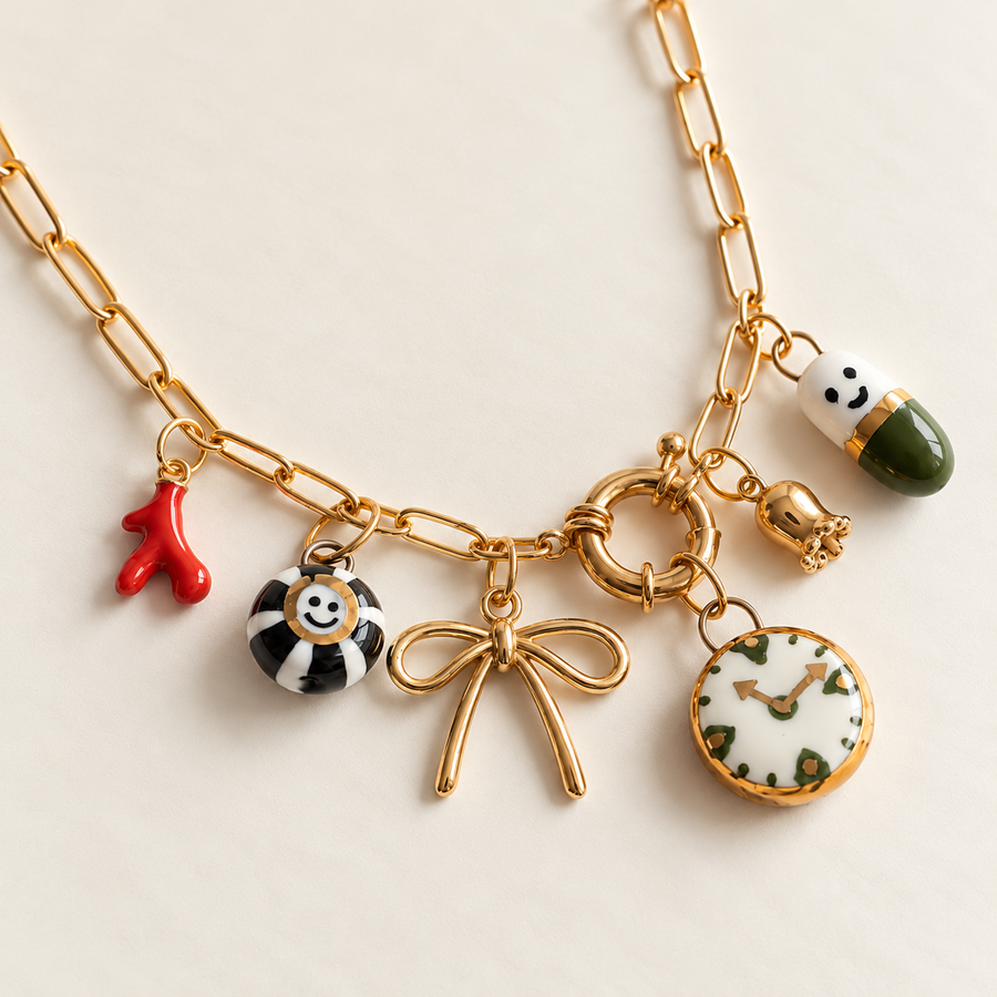 A gold chain necklace with multiple charms, including a red coral-like charm, a black-and-white striped smiley charm, a gold bow charm, a round floral clock charm, and a green-and-white capsule charm, displayed on a light beige background.