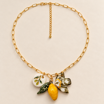 A gold chain necklace with paperclip-style links and a cluster of ceramic charms, including a yellow lemon, green leaves, a white flower with green leaves, and a small white oval charm, displayed on a light beige background.