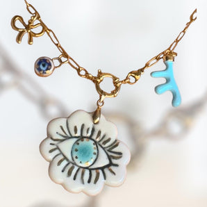 A handmade gold chain necklace with unique charms: a turquoise abstract hand, a blue and white evil eye, a gold bow, and a large ceramic flower pendant featuring an eye design with a turquoise center.