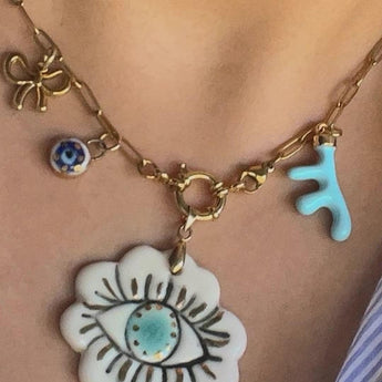 A handmade gold chain necklace with unique charms: a turquoise abstract hand, a blue and white evil eye, a gold bow, and a large ceramic flower pendant featuring an eye design with a turquoise center.