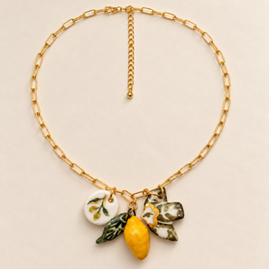 A gold chain necklace with paperclip-style links and a cluster of ceramic charms, including a yellow lemon, green leaves, a white flower with green leaves, and a small white oval charm, displayed on a light beige background.
