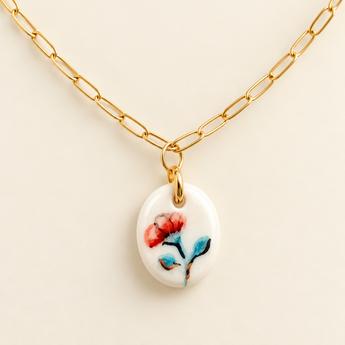 A gold paperclip chain necklace with an oval white pendant featuring a painted red and blue flower, displayed on a light beige background.
