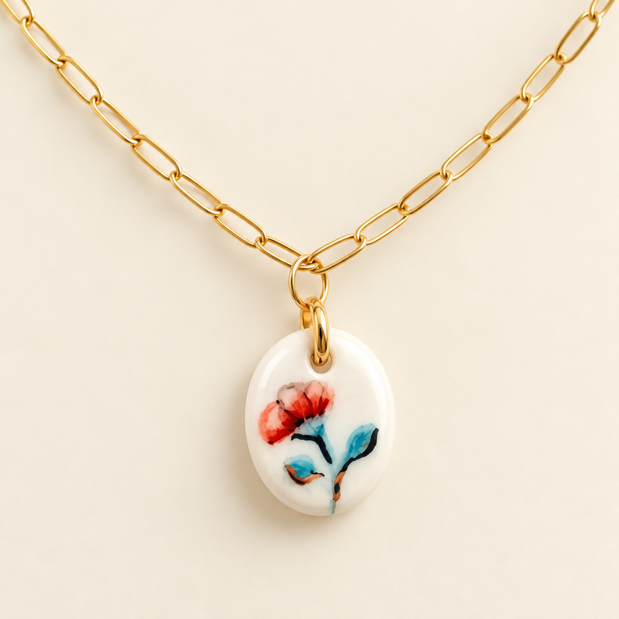 A gold paperclip chain necklace with an oval white pendant featuring a painted red and blue flower, displayed on a light beige background.