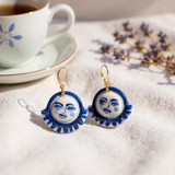 Celestial Gaze Porcelain Earrings alongside a cup of tea, highlighting these unique statement earrings with cobalt blue moon face design and gold accents.