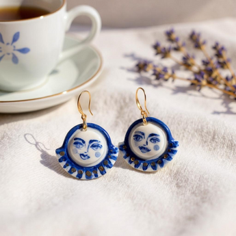 Celestial Gaze Porcelain Earrings alongside a cup of tea, highlighting these unique statement earrings with cobalt blue moon face design and gold accents.