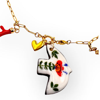 A handmade gold chain bracelet featuring unique charms: a ceramic bird-shaped pendant with hand-painted red and green floral details, a red coral-shaped charm, a small gold heart, and a gold seashell. The bracelet has a lightweight and artistic