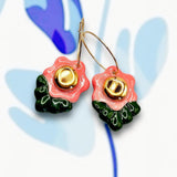 A pair of handmade porcelain flower earrings with coral pink petals, deep green leaves, and gold detailing, attached to gold hoops, displayed on a colorful background.