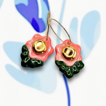 A pair of handmade porcelain flower earrings with coral pink petals, deep green leaves, and gold detailing, attached to gold hoops, displayed on a colorful background.